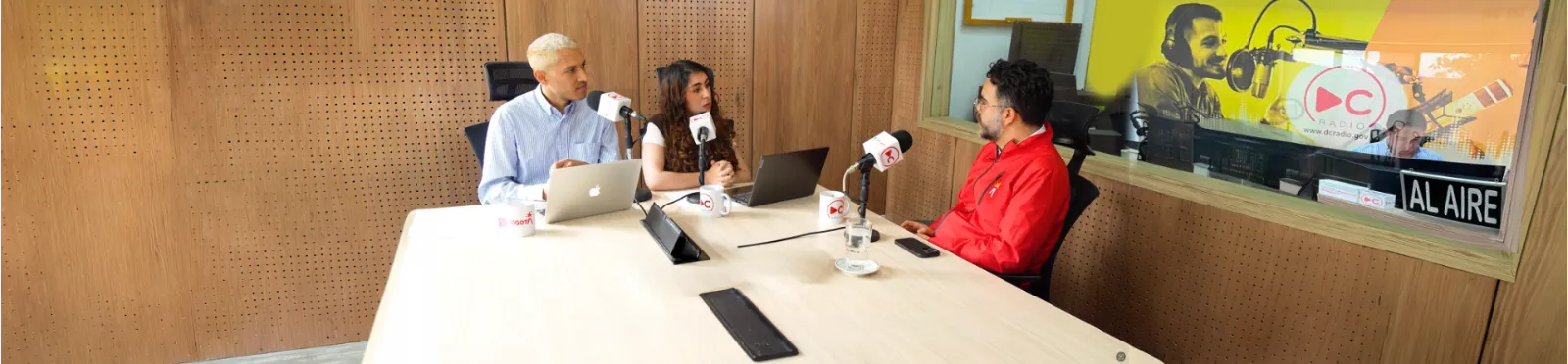 Tres personas conversan en el estudio de DC Radio: dos locutores frente a micrófonos y un invitado con chaqueta roja. Al fondo, la cabina con el letrero "Al aire".